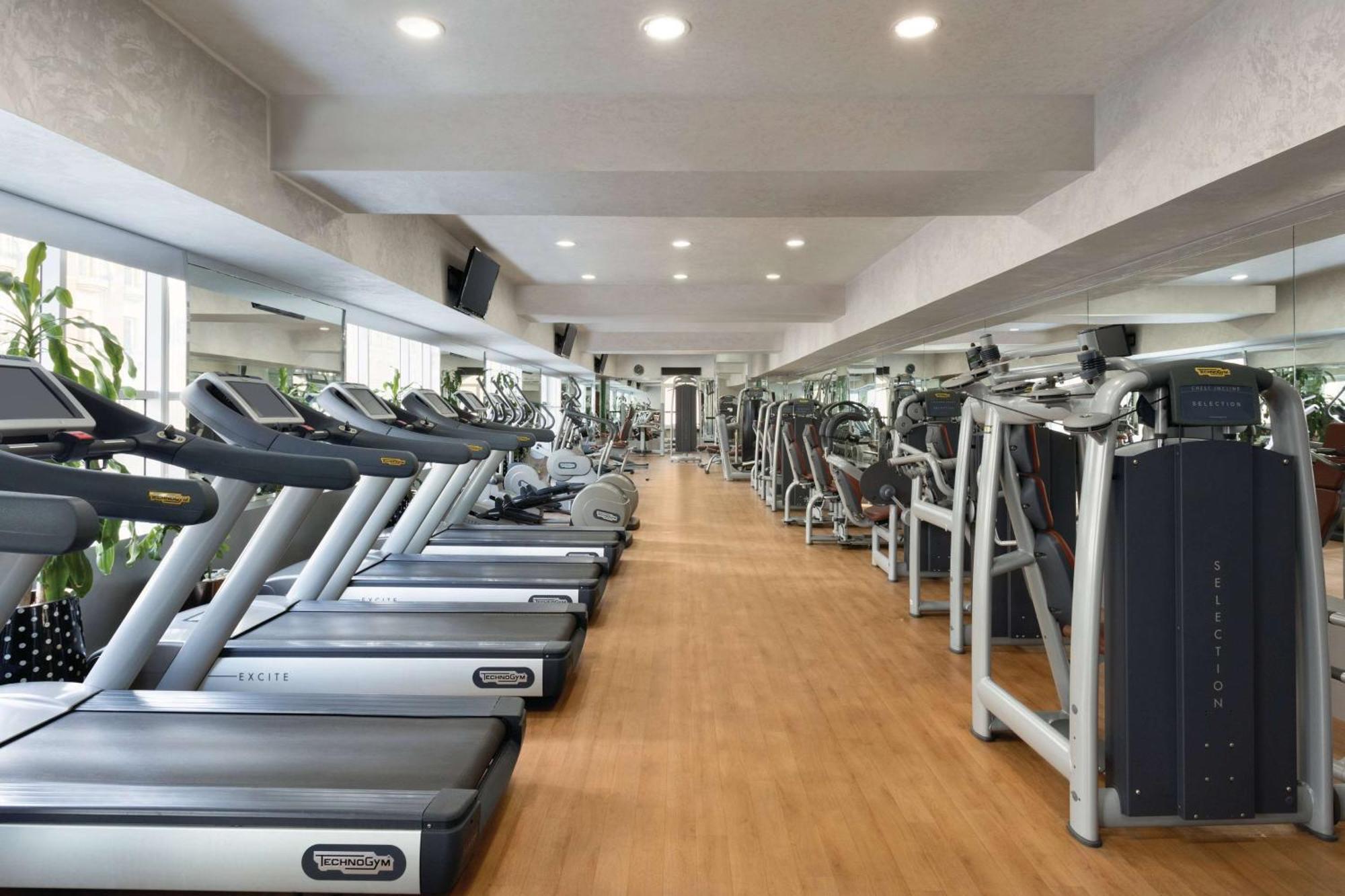 Grand Regency Doha, Trademark Collection By Wyndham Bagian luar foto The photo shows the interior of a gym or fitness center. It features a row of modern treadmills lined up against a wall, with a polished wooden floor. The space is brightly lit with recessed ceiling lights, and there are large mirrors on the walls, e