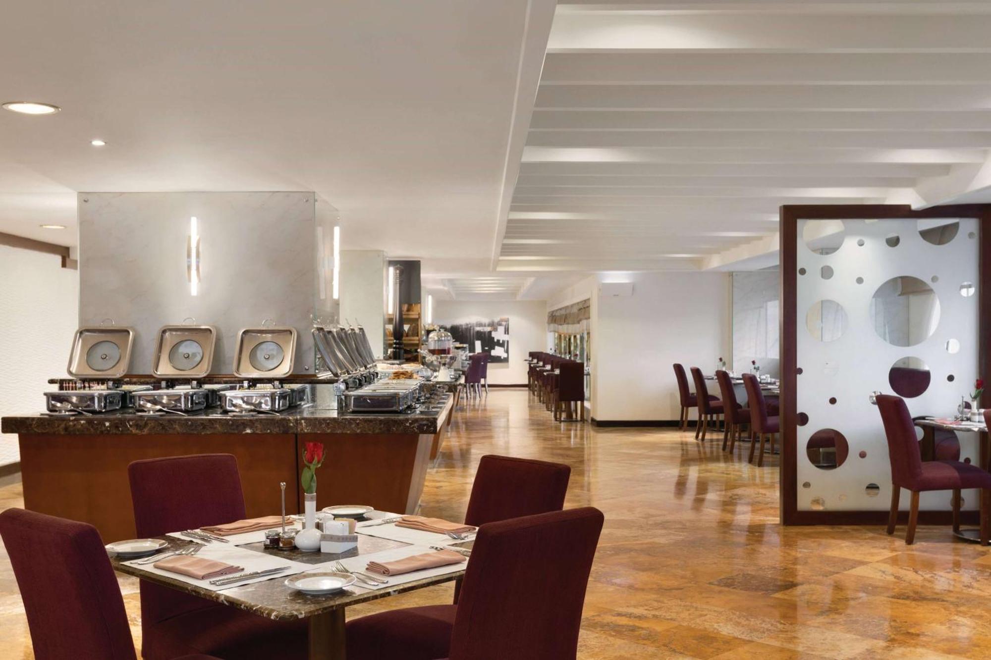 Grand Regency Doha, Trademark Collection By Wyndham Bagian luar foto The photo shows a modern restaurant interior. There are several tables with maroon chairs arranged throughout the space. One table is set with a small vase containing a flower and dining utensils. Along one side, there is a buffet area with a sleek c