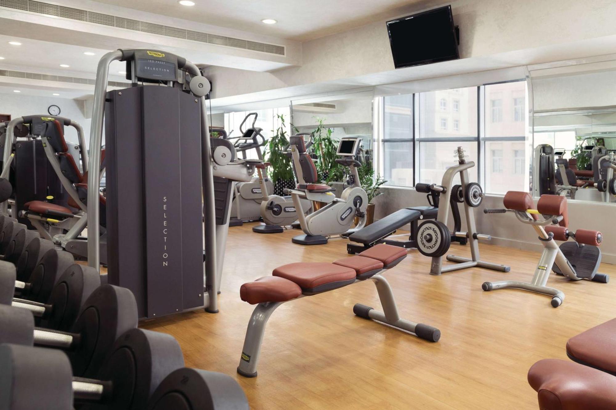 Grand Regency Doha, Trademark Collection By Wyndham Bagian luar foto The photo shows a fitness gym with various exercise equipment. There are weight machines, benches, and cardio machines such as stationary bikes and treadmills. The space has large windows allowing natural light to enter, and there are mirrors on the 