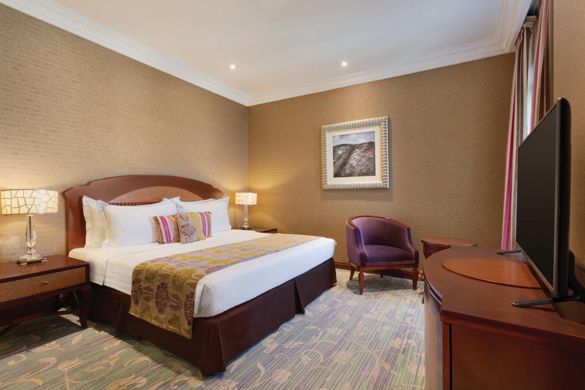 Grand Regency Doha, Trademark Collection By Wyndham Bagian luar foto The image shows a hotel room that features a cozy and elegant design. There's a large bed with white sheets and colorful decorative pillows. Next to the bed, there are nightstands with lamps. A comfortable armchair is positioned in the corner, and a 
