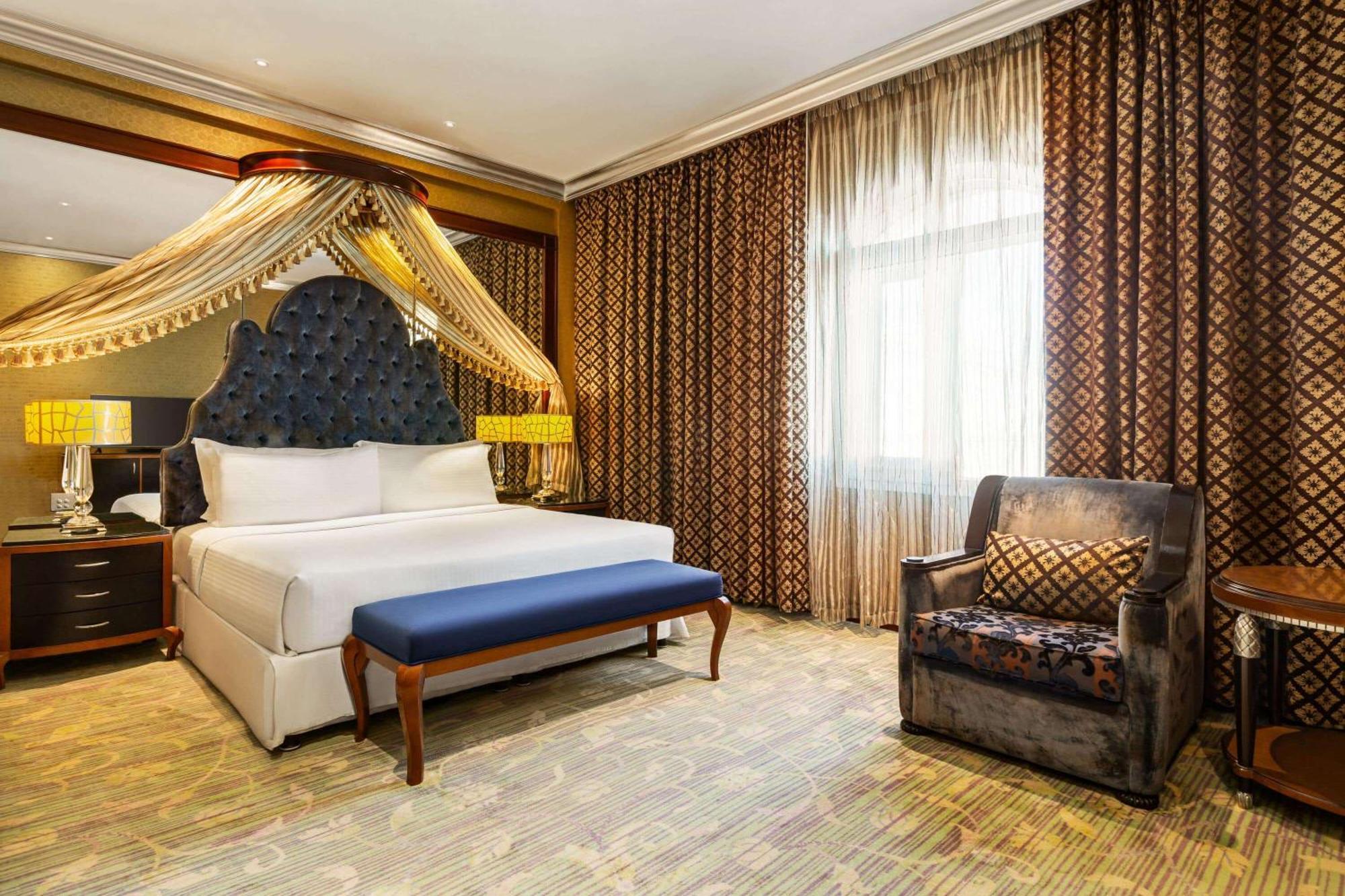 Grand Regency Doha, Trademark Collection By Wyndham Bagian luar foto The photo shows a luxurious hotel room featuring a large bed with an intricately designed headboard and elegant bedding. Above the bed, there are decorative drapes that add a touch of sophistication. In front of the bed, there is a blue bench. To the