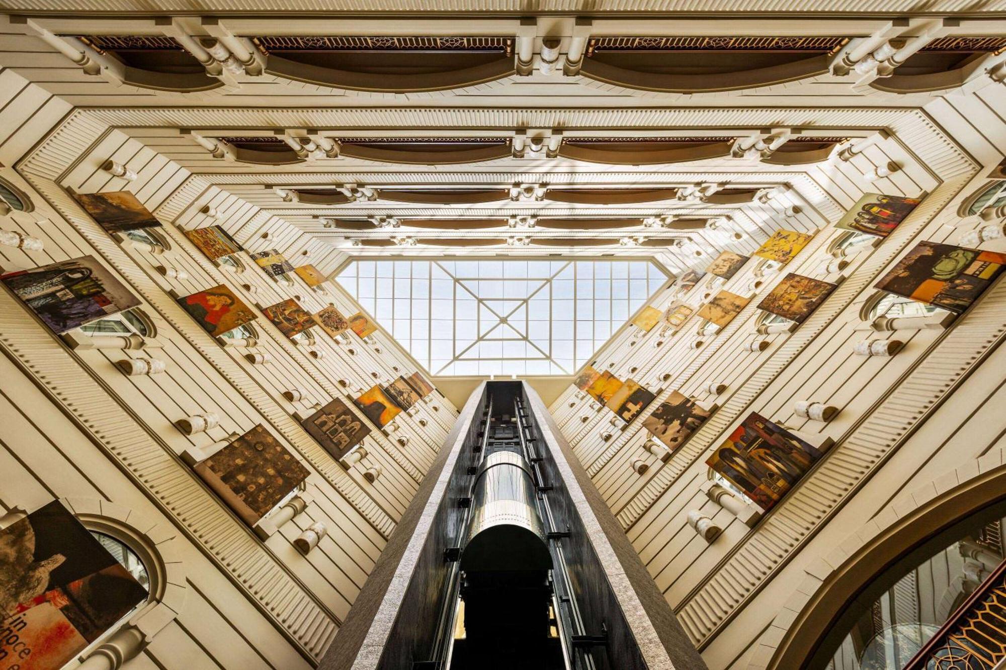Grand Regency Doha, Trademark Collection By Wyndham Bagian luar foto The photo shows an interior view of a building, captured from a low angle looking upwards. The architectural design features a tall, open space with walls adorned with various artworks. In the center, there is a glass ceiling that allows natural ligh