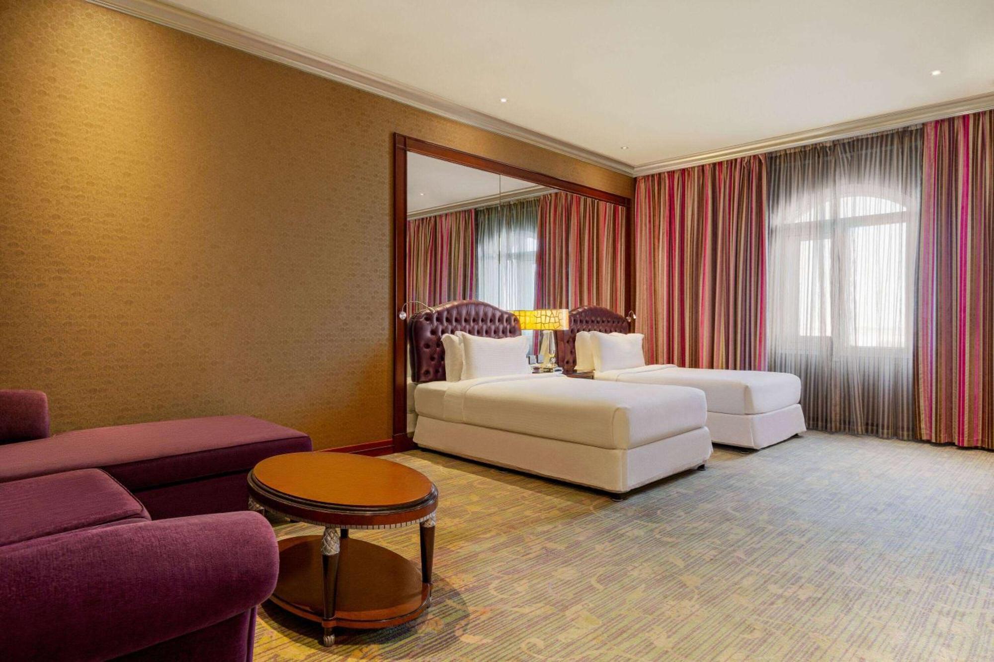 Grand Regency Doha, Trademark Collection By Wyndham Bagian luar foto The photo shows a hotel room featuring two beds with white bedding, a large mirror on one wall, and a window dressed with sheer and curtain panels. There is a small round table in the center of the room and a plush purple armchair nearby. The overall