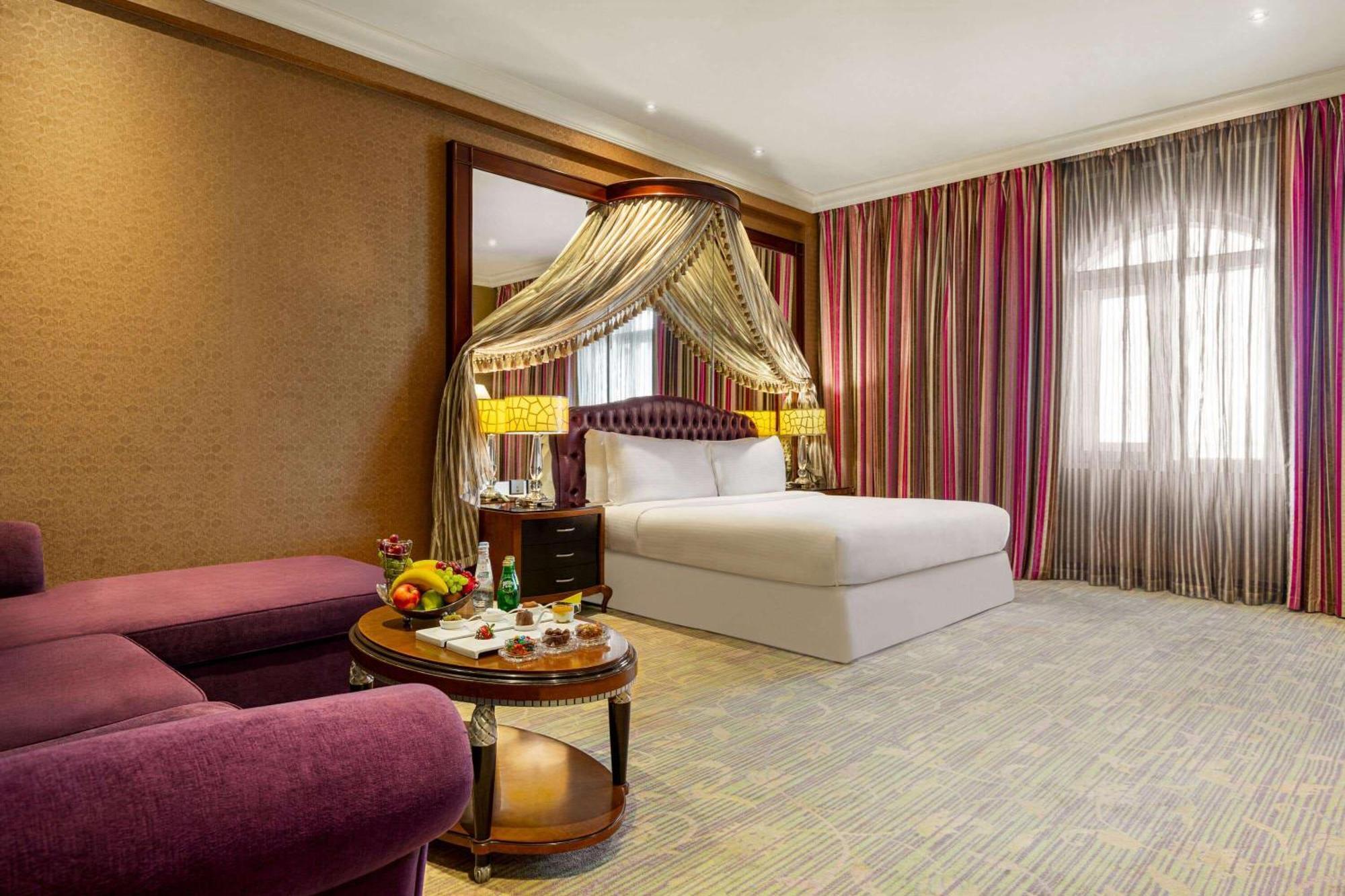 Grand Regency Doha, Trademark Collection By Wyndham Bagian luar foto The photo shows a luxurious hotel room featuring a king-sized bed with a decorative canopy. The room is elegantly designed with a mix of rich colors and textures. To the left, there's a plush purple seating area with a small round table that displays
