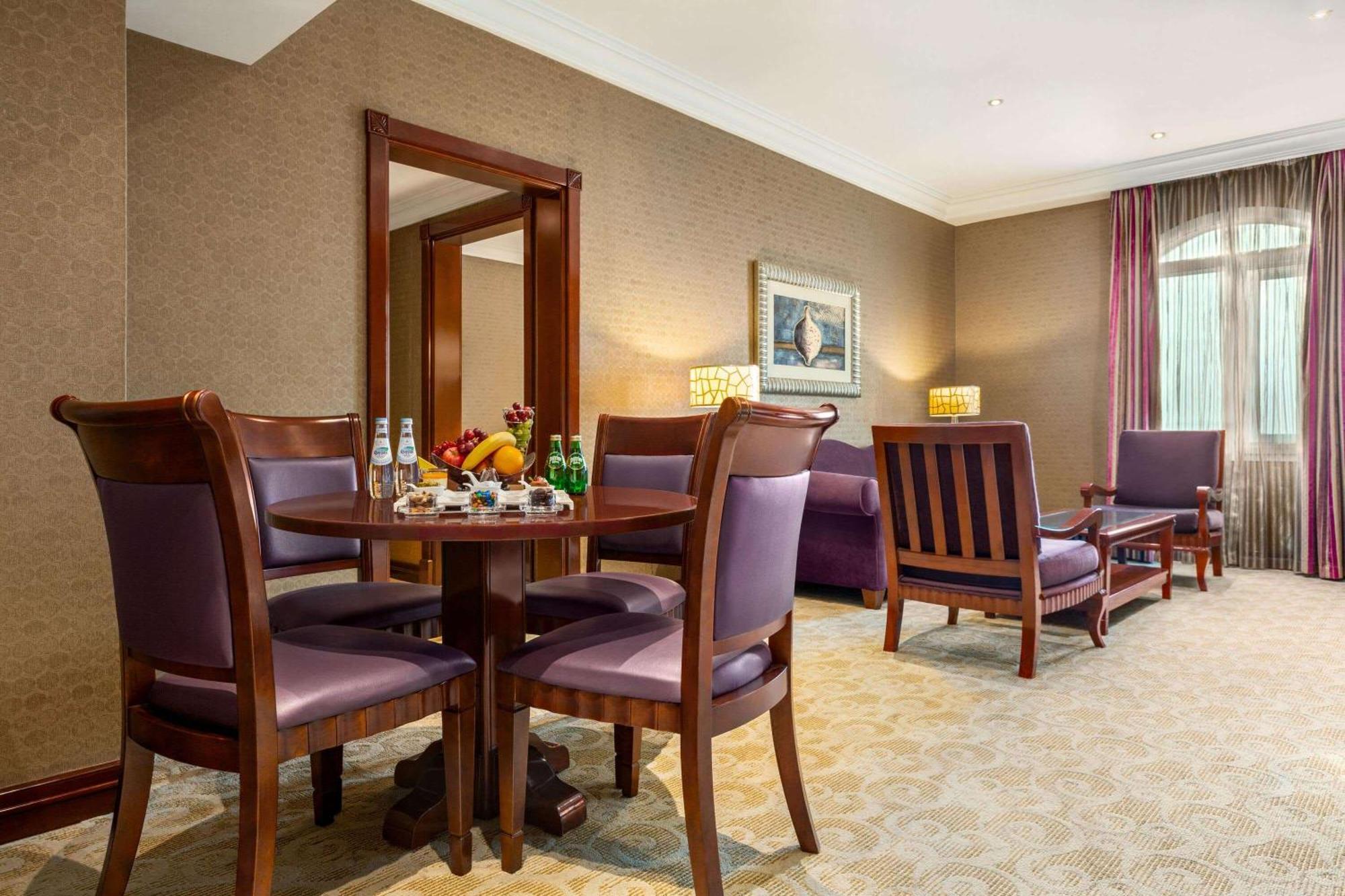 Grand Regency Doha, Trademark Collection By Wyndham Bagian luar foto The photo depicts a well-furnished hotel room or lounge area. In the foreground, there is a round table surrounded by four purple upholstered chairs, with a selection of fruits and beverages on the table. To the right, there are two wooden armchairs,
